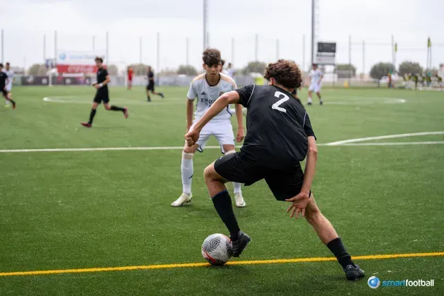 Soccer player in black jersey dribbles, facing opponent in white, on green field.