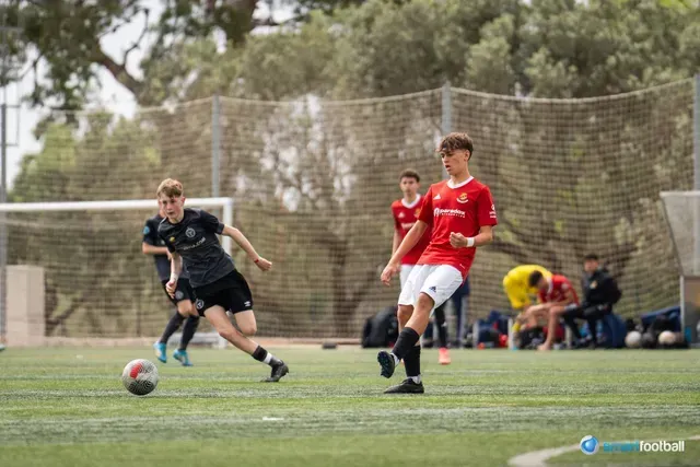 Soccer players on a green field. A player in red runs with the ball, another in black tries to intercept.