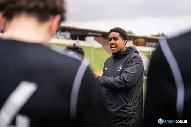 Soccer coach giving instructions to his team on the field, wearing a black jacket.