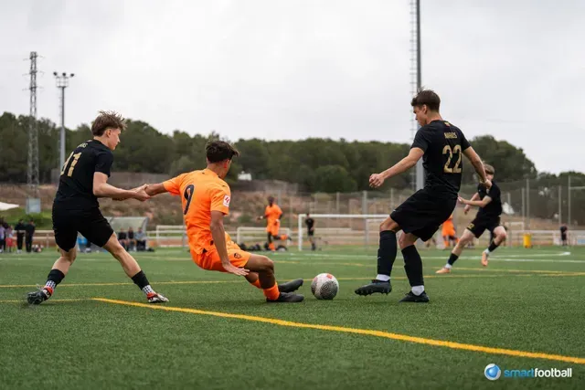 Soccer players in action: Orange jersey player slides to block ball, two black-jerseyed players attempt to intercept. 