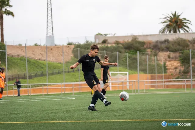 Soccer player in black kicks ball on green field. Others in background.