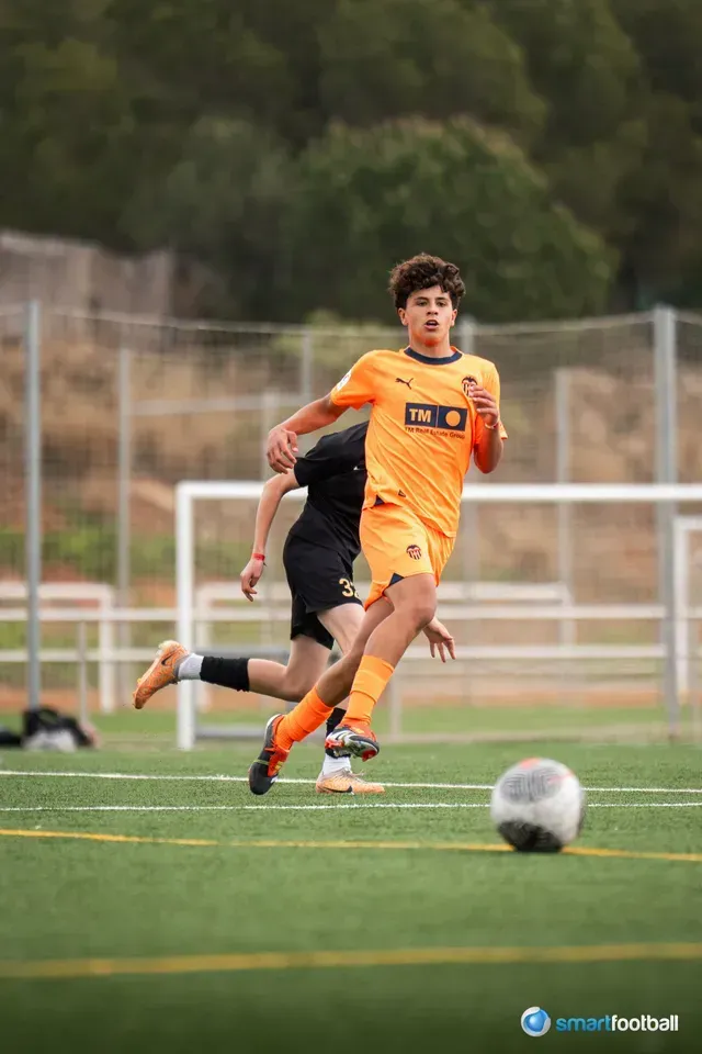 Soccer player in orange running towards the camera, ball in foreground, opponent behind. Green field, outdoor setting.