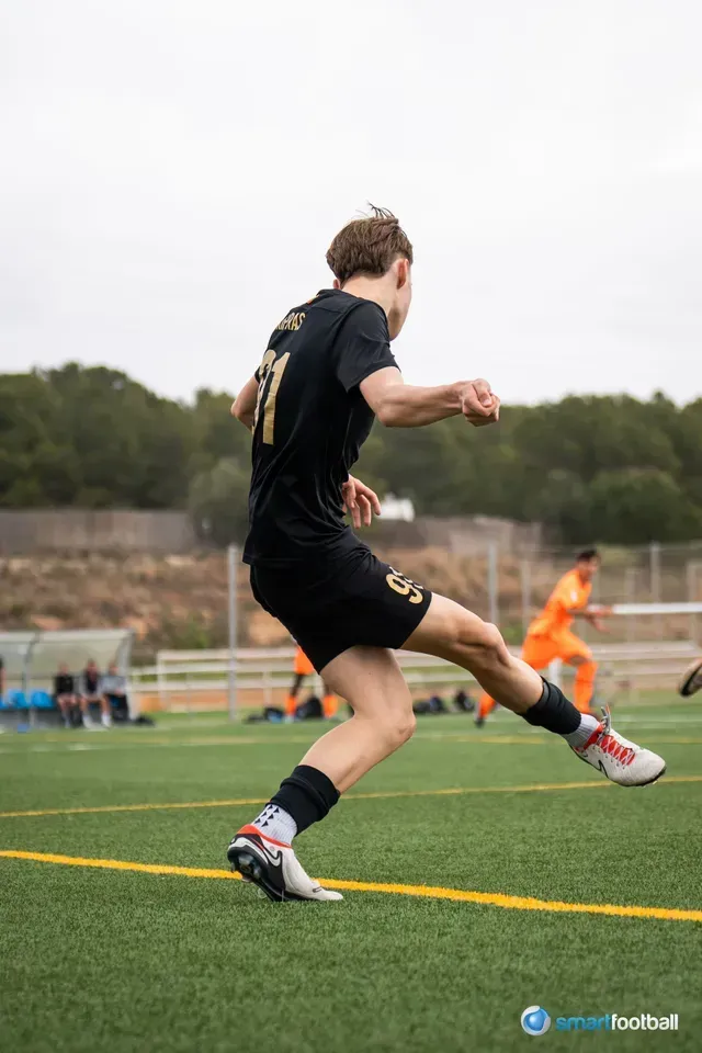 Soccer player in black uniform kicks the ball on a green field. Other players in orange uniforms in background.