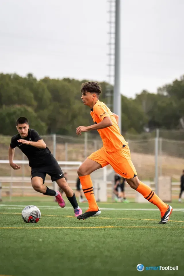 Soccer players on a field. One in orange runs with the ball, the other in black pursues. Cloudy sky.