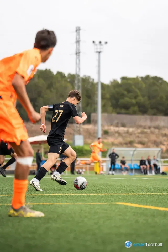 Soccer player in black jersey with ball, running on field during game. Orange jersey opponent nearby.