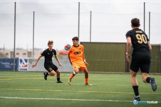 Soccer players in action on a green field. An orange-clad player heads the ball, while two black-clad players give chase.