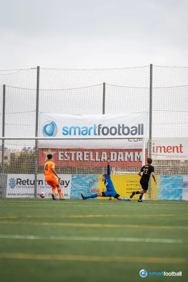 Soccer game: Player kicks ball toward goal. Goalie dives. Another player runs towards the goal.
