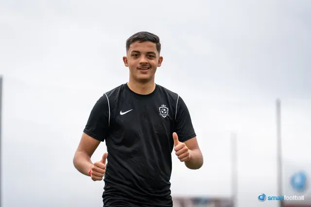 Youth soccer players in action on a green field. Two players sprint, one with the ball, wearing blue and red uniforms.