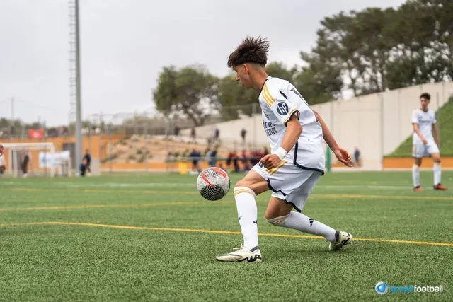 Soccer player in white uniform dribbles a ball on a green field, with others in the background.