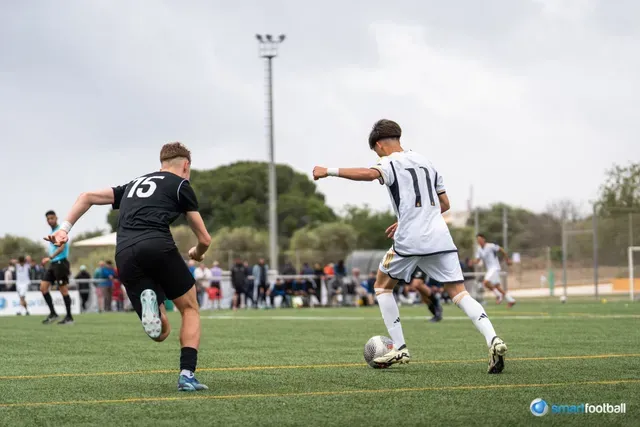 Soccer players in action on a green field; one in black, chasing the other with the ball in a white uniform.