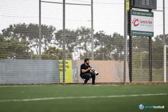 Photographer squats on a soccer field, holding a camera.  Background includes a fence, sign, and scoreboard.