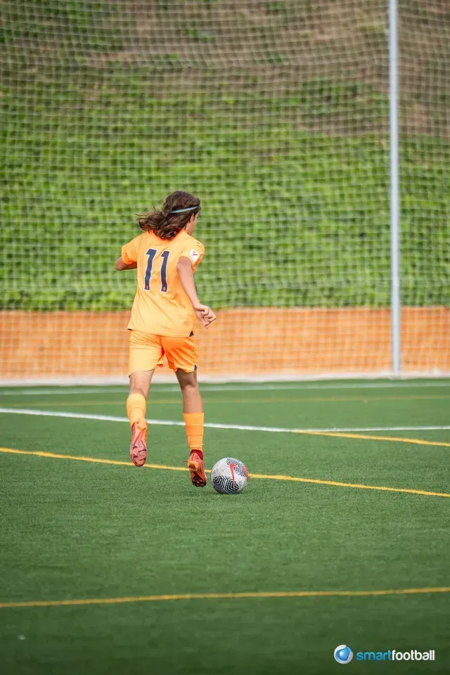 Soccer player in orange uniform dribbling the ball on a green field, with a net and trees in the background.