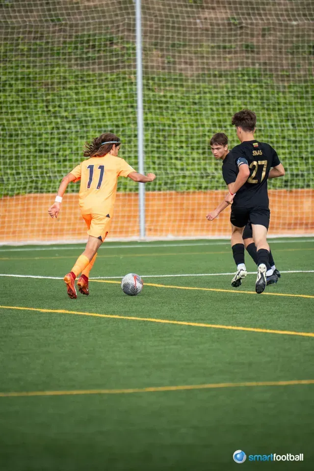 Soccer players in action on a green field. A player in orange kicks the ball, pursued by two in black.