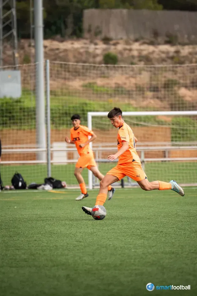 Soccer player in orange uniform kicking ball on a green field. Another player watches in the background.