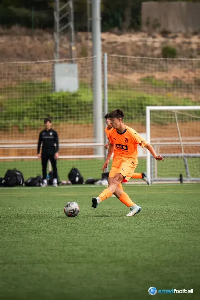 Soccer player in orange uniform looks up with hands clasped, celebrating on a field with teammates.