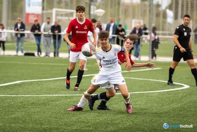 Soccer players on field; one in white passes while two others attempt to intercept. Spectators watch in the background.