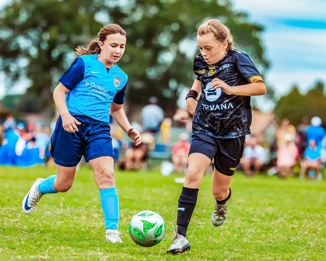 Two young girls playing soccer on a green field. One in blue, one in black, chasing the ball.