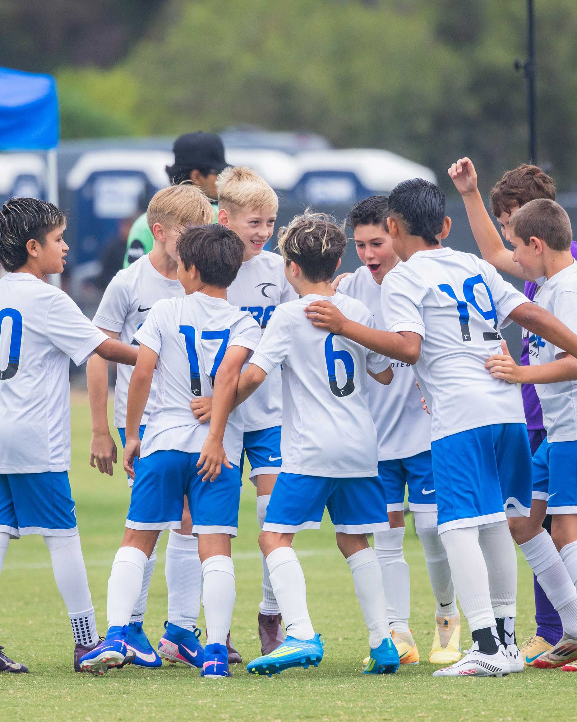 Youth soccer team in white and blue huddles on the field, arms around each other in celebration.