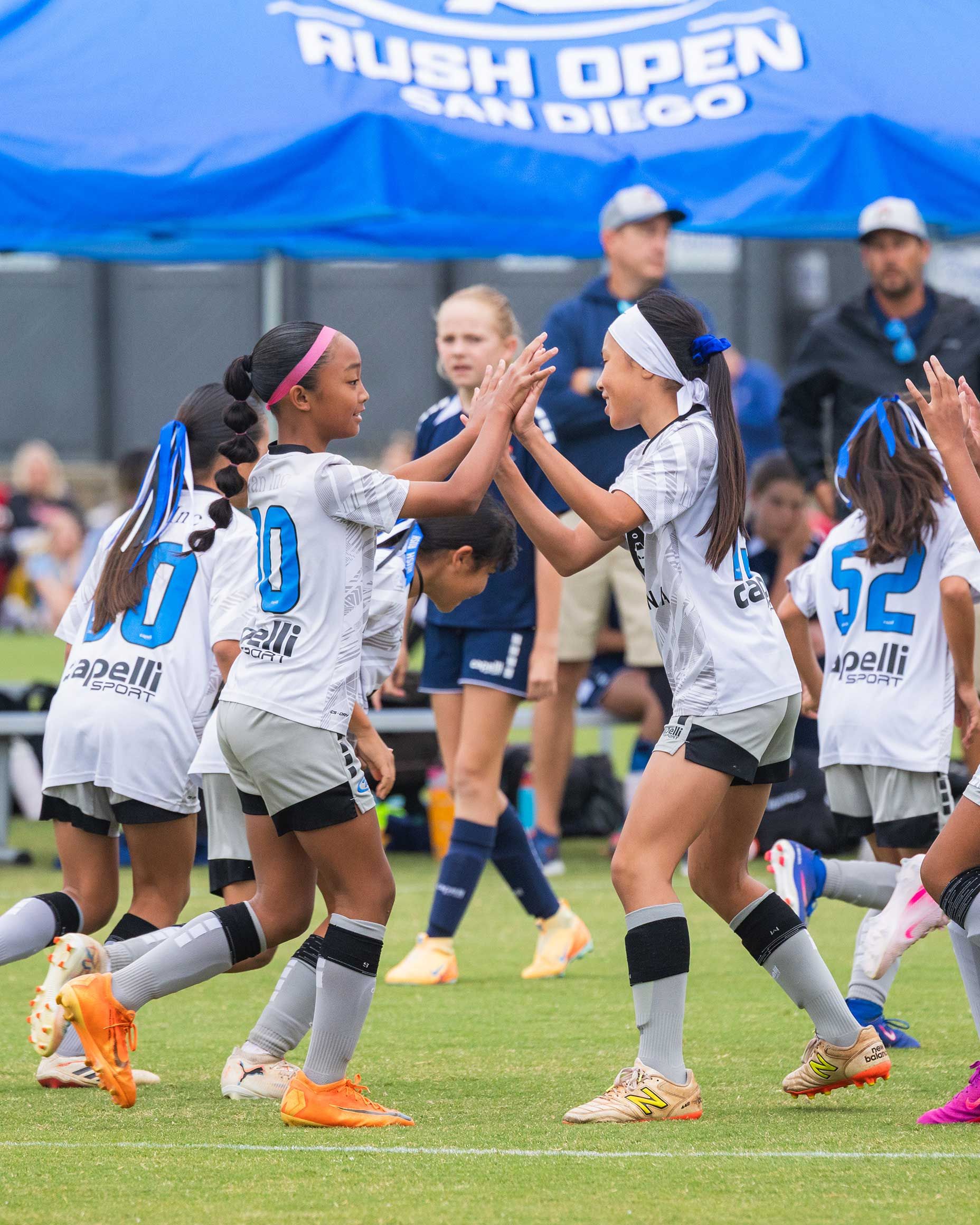 Youth soccer players in blue and white uniforms high-five on a rainy field during a Rush Open event
