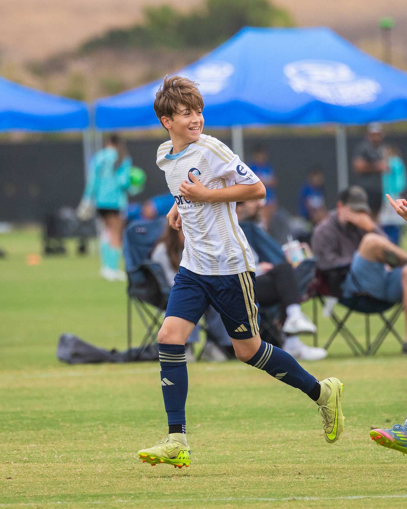 Soccer player in white jersey and navy shorts running on a grassy field with blue tents in the background