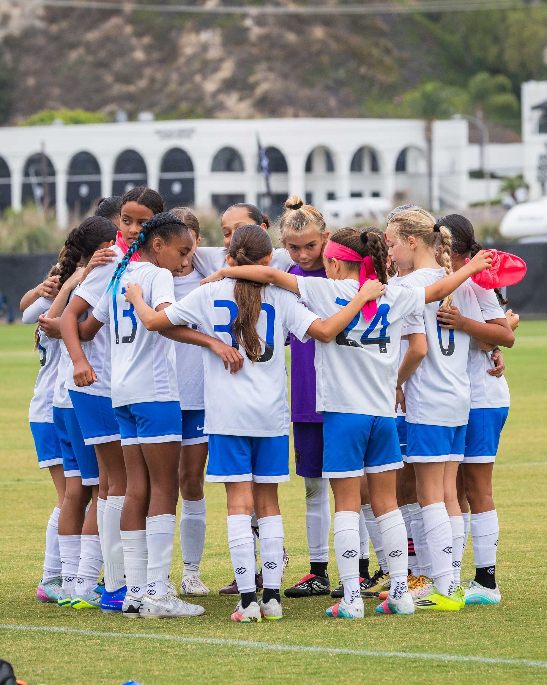 Soccer team in white and blue uniforms huddles on a grassy field before a match