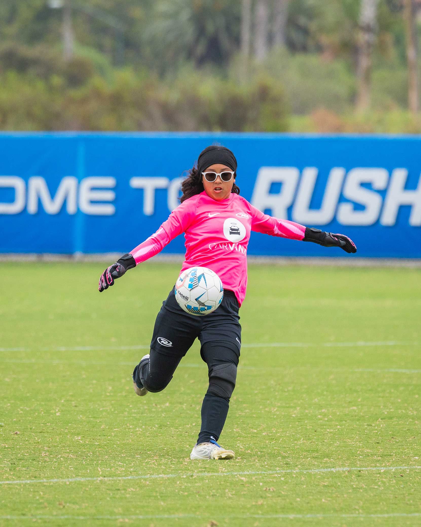 Soccer goalkeeper in pink jersey and black pants kicking a ball on a grassy field
