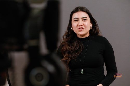 Donna che parla, indossa un top nero, ha i capelli scuri, una collana e uno sfondo grigio.