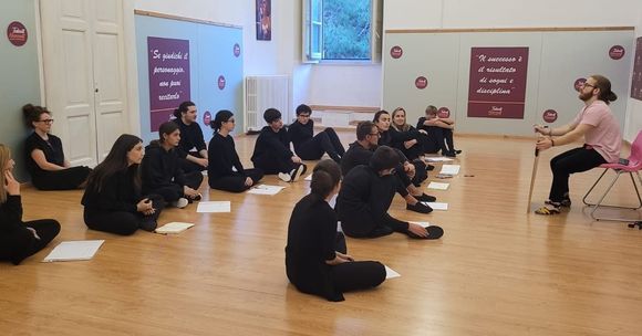 Un gruppo di persone vestite di nero è seduto sul pavimento e ascolta una persona seduta con un bastone.