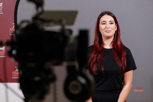 Una donna con i capelli tinti di rosso è in piedi davanti a una telecamera e a uno sfondo rosso, sorridendo leggermente.