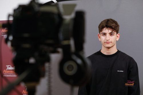 Un giovane uomo con una camicia nera è in piedi vicino a una telecamera, pronto a filmare su uno sfondo grigio.