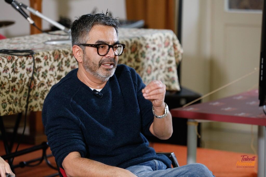 Un uomo con gli occhiali gesticola mentre parla, seduto vicino a un tavolo con una tovaglia a fantasia.