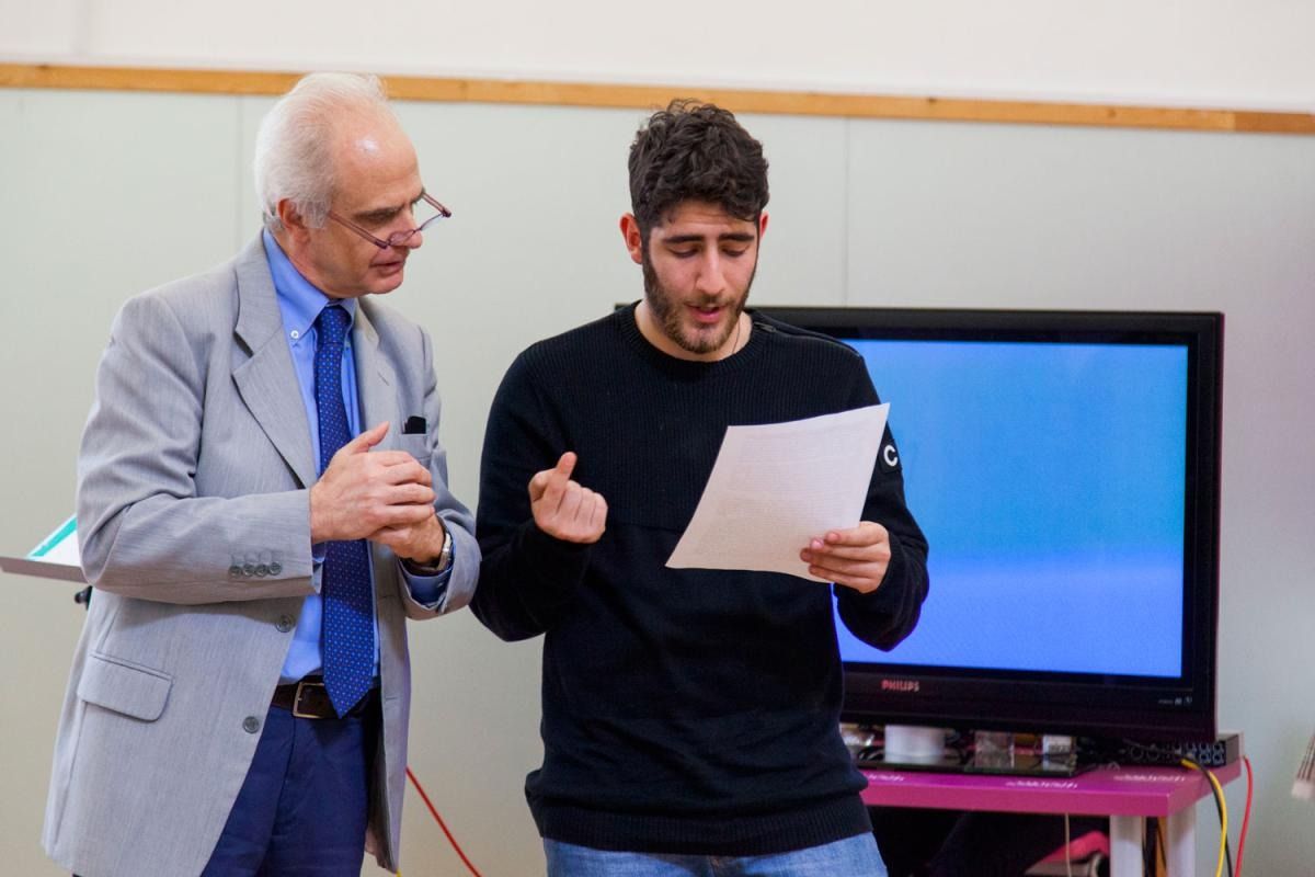 Un uomo anziano in giacca e cravatta osserva un uomo più giovane leggere un giornale, con la TV sullo sfondo.