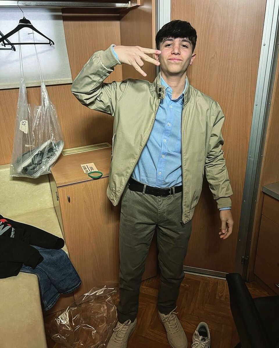 Un giovane con giacca e pantaloni color cuoio e camicia blu fa un gesto con la mano in una stanza con pannelli di legno.