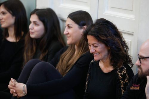 Persone sedute per terra, sorridenti. Una donna con un top nero, altre in abiti scuri. Pareti bianche.