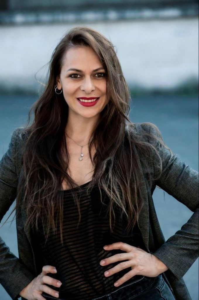 Donna con lunghi capelli castani, mani sui fianchi, indossa un blazer e un top nero, sorridente, all'aperto.