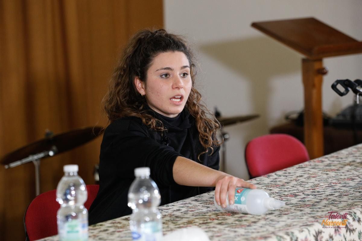 Donna che parla seduta a un tavolo, con in mano una bottiglia d'acqua. Capelli scuri, top nero, in un interno con un podio e delle bottiglie.