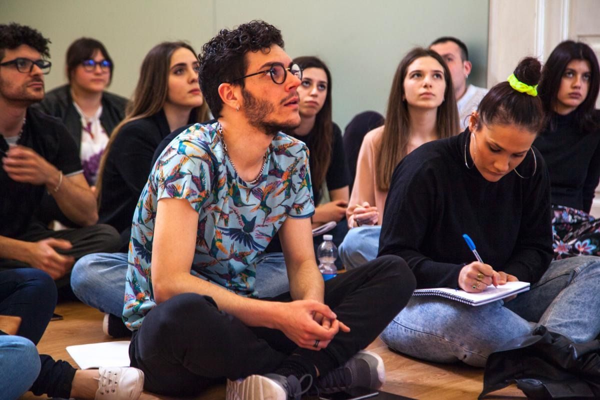 Un gruppo di persone sedute sul pavimento, alcune guardano verso l'alto, altre scrivono, in una stanza.