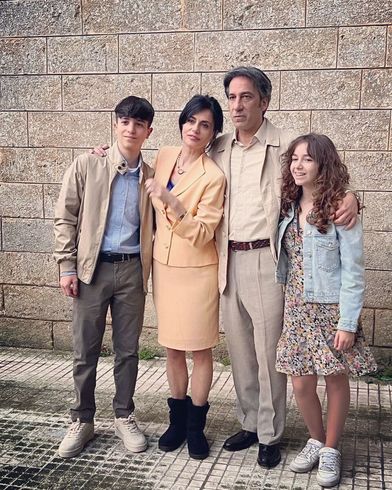 Una famiglia di quattro persone posa davanti a un muro di pietra. Un uomo e una donna abbracciano due giovani adulti, tutti vestiti in modo casual.