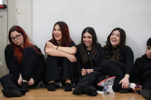 Cinque persone sono sedute e ridono contro un muro, tutte vestite di nero.