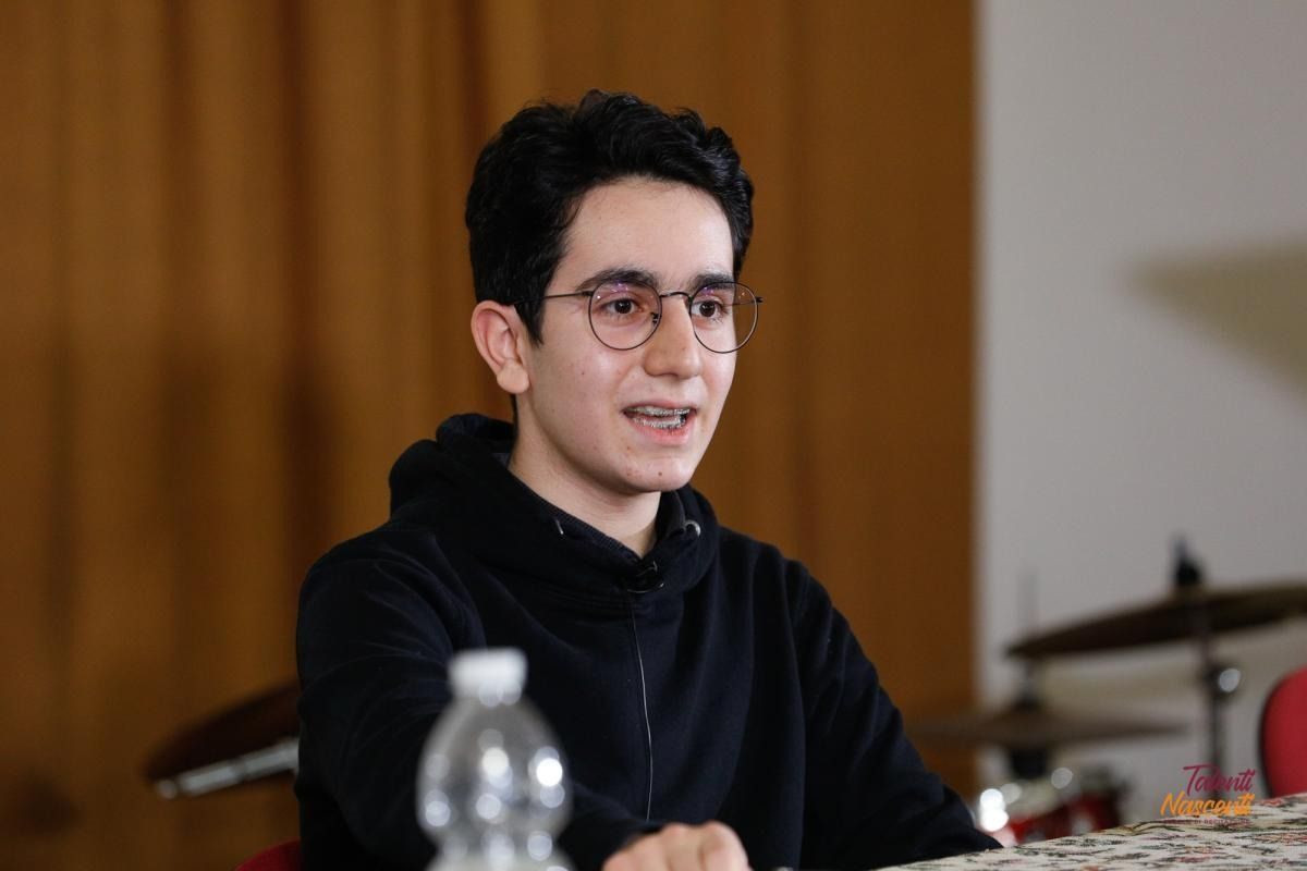 Giovane con gli occhiali, sorridente, con una felpa nera. Seduto davanti a uno sfondo neutro, possibilmente al chiuso.