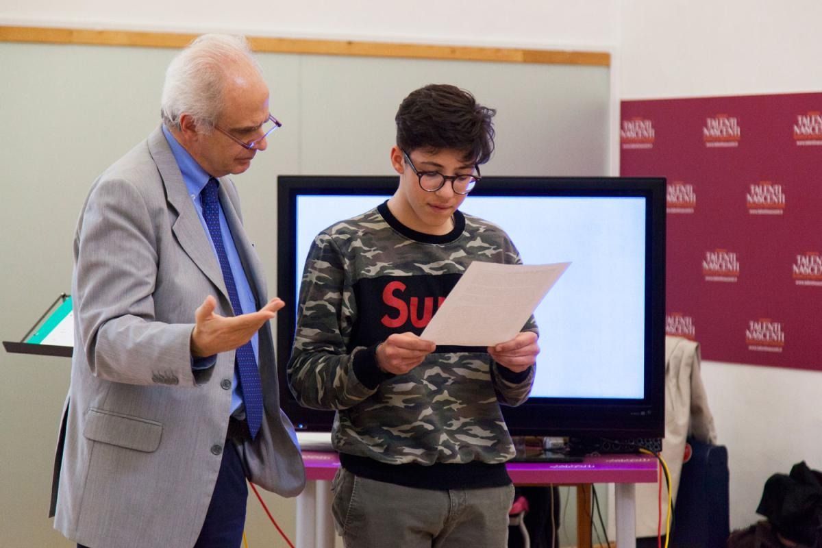 Un uomo in giacca e cravatta indica un adolescente che sta leggendo un giornale; si trovano in una stanza con uno schermo e un tavolo color magenta.