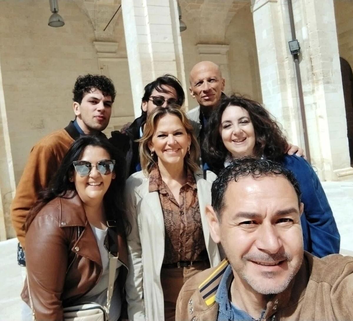 Un gruppo di sette persone sorridenti, in posa per un selfie in un edificio con colonne ad arco.
