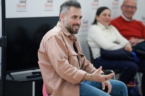 Un uomo con una giacca marrone gesticola mentre parla, seduto davanti a un monitor e a due persone.