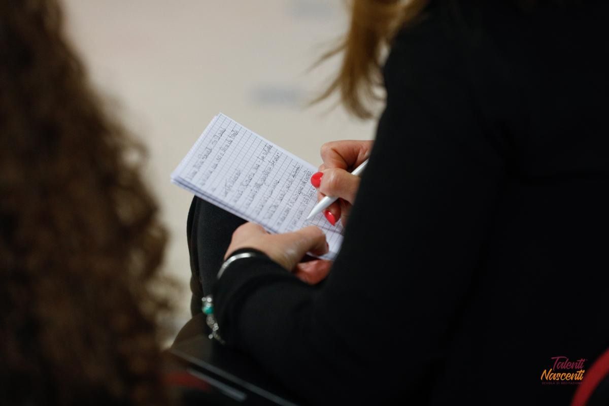 Persona che scrive su un blocco note, smalto rosso sulle unghie, maniche nere.