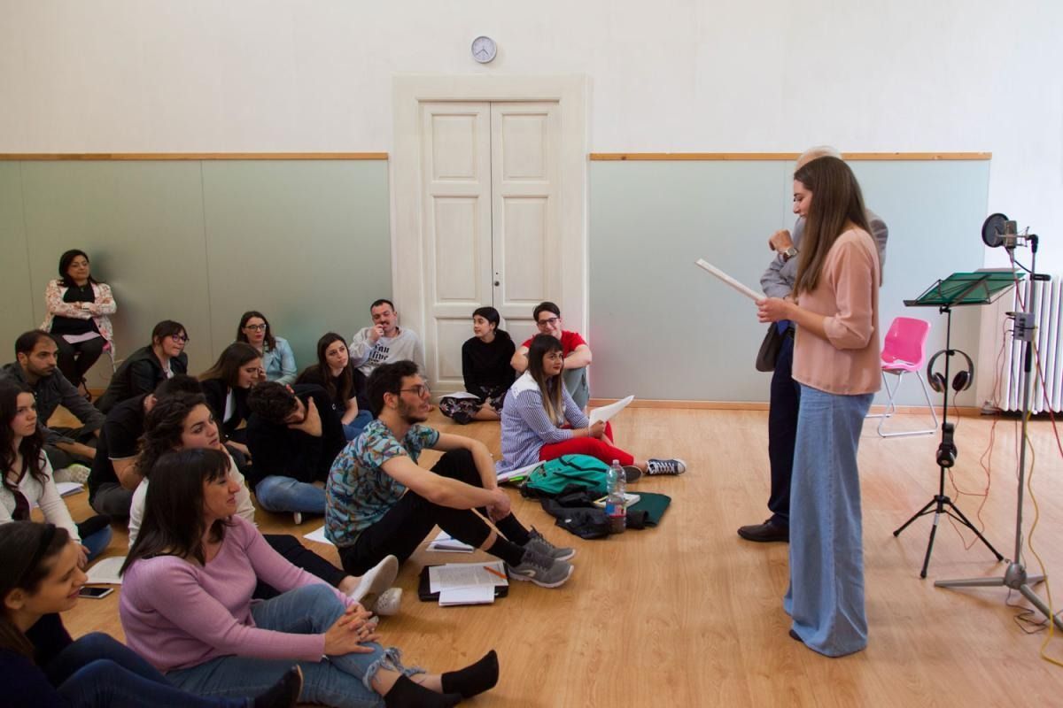 Una giovane donna si esibisce in una stanza con un pubblico seduto, probabilmente durante una prova o un workshop.