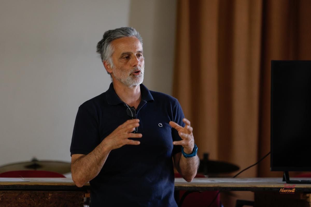 Un uomo con barba e capelli grigi gesticola mentre parla, probabilmente sta tenendo una presentazione.