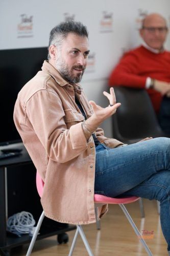 L'uomo gesticola mentre è seduto, indossa una camicia marrone chiaro, jeans ed è seduto su una sedia rosa, in un ambiente interno.