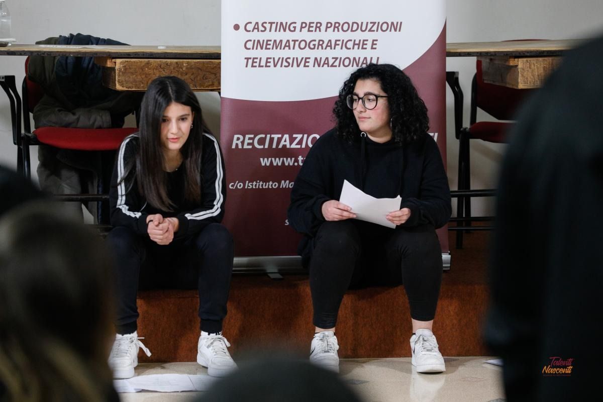 Due giovani donne sedute leggono copioni in una stanza con uno striscione che dice