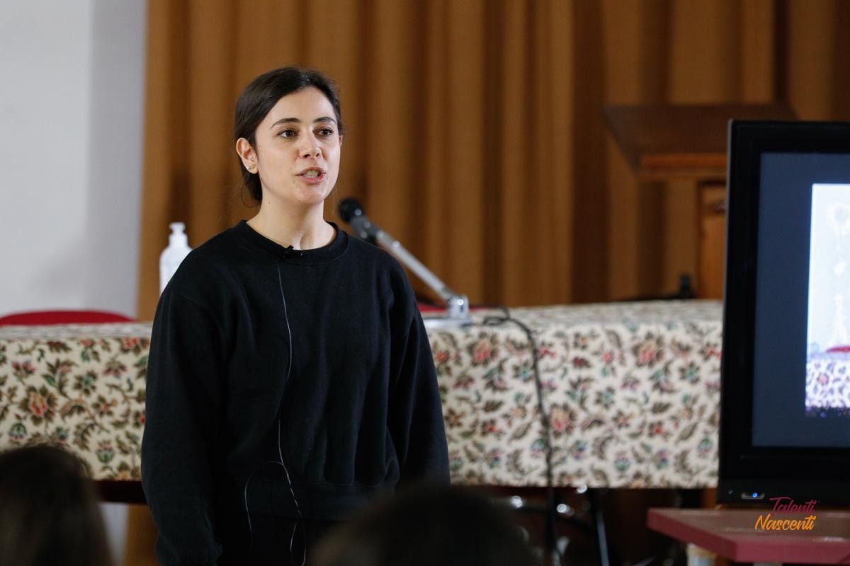 Una donna parla durante una presentazione, usando un microfono. È in piedi davanti a uno schermo con una tovaglia a motivi floreali alle sue spalle.