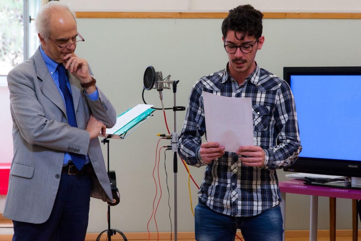 Un uomo legge un foglio di carta, di fronte al microfono, mentre un altro uomo osserva. All'interno dello studio.